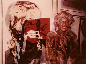 A Photograph of Marsha P. Johnson Standing in Front of a 1975 Portrait of Her by Andy Warhol