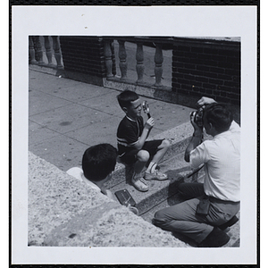 A boy sitting on the stairs and holding a magnifying glass over his eye as a photographer takes pictures during a Boys' Club Freckle King Contest