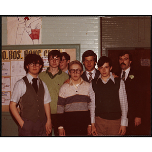 Six young men and a man with a carnation on his lapel pose together in front of a bulletin board during a Boys' Club St. Patrick's Day event at the South Boston Clubhouse