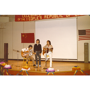 Three Chinese men sing and play guitar onstage at the 30th anniversary celebration of the People's Republic of China held in the Josiah Quincy School auditorium