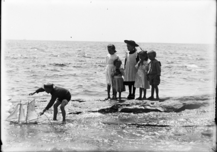A young boy sails his toy yacht along the shore as a group of children watch.