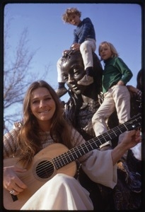 Judy Collins seated at the base of a statue, playing guitar for children