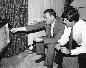 Mayor Raymond L. Flynn tuning a television while seated on a bed with John Kerry