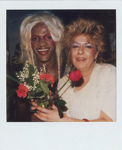 A Photograph of Marsha P. Johnson With Blonde Hair and Red Eyeshadow, Smiling with an Unknown Person Holding a Flower Bouquet