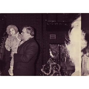 Mayor Menino with a child in a fur-lined hood in front a Villa Victoria house decorated for Christmas.