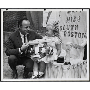 The Little Sister Contest winner sitting with a doll as a man spreads her sash for the camera