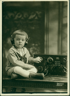 Portrait of a young boy, seated, facing front, listening to a radio,location unknown, undated