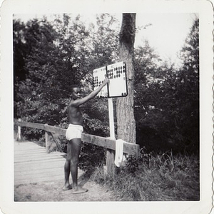 A boy in swimming trunks poses in front of a sign at Breezy Meadows Camp