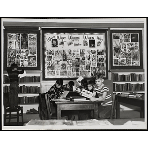 A group of boys work at a table below a "Who What Where When Why" triptych collage in the South Boston Clubhouse library. Photo caption reads: "The Library Project: 'Who-What-When-Where-Why' Report outlining a quiz contest, which was held in the library of the Charles Hayden Memorial Clubhouse"