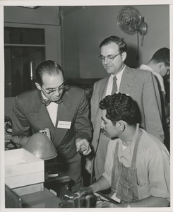 Unidentified man demonstrating eyeglass lens production for two onlookers