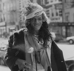 A Photograph of Marsha P. Johnson Posing in the Street Wearing a Black Jacket with a Sweater Tied Around Her Neck