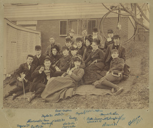 Group of students sitting on a blanket on the grass with a building and a penny-farthing bicycle in the background