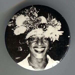A Pin of Marsha P. Johnson Wearing an Elaborate Flower Headpiece and Smiling