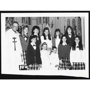 A boy and a girl in their communion outfits posing with a priest, girls in school uniform, and two guests