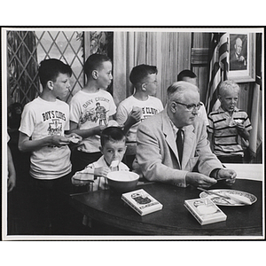 Six boys eating stand behind radio host and WBZ-TV presenter Bob Emery seated at a desk