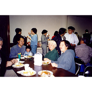 Association members and their families eat Thanksgiving dinner, with people standing in line to get food in the background