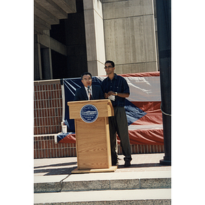 Two men stand at a lectern in City Hall Plaza