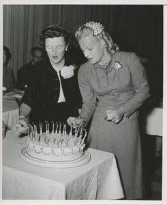 Margaret Milbank Bogert and another woman light candles on a cake at the Institute for the Crippled and Disabled's 35th anniversary Red Cross luncheon