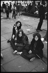 Three young women sit on the sidewalk, observing the Moratorium protests