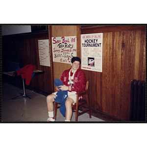A young man in a warm-up jacket holds a pair of pants and looks at the camera, while sitting in front of a wall with posters announcing "Super Bowl '88 Teen Party" and "News America Cup Hockey Tournament," at the Charlestown Boys & Girls Clubhouse