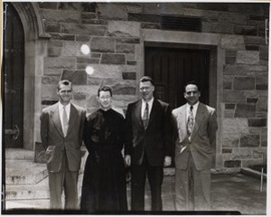 Four men standing outside building at Boston College, one is a member of the clergy
