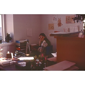 Female employee wearing eyeglasses seated at a desk, at La Alianza Hispana, Roxbury, Mass