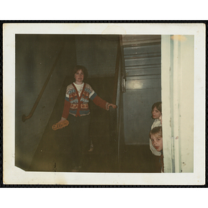 A woman looks at the camera while walking down the stairs with a nameplate reading "Ellen" in her hand while several children stand behind a wall at the South Boston Boys' Club