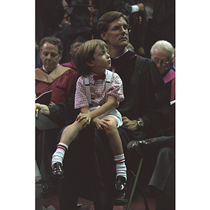 Child sits on faculty member's lap at commencement