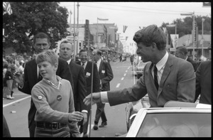 Robert F. Kennedy (left) seated in an open car at the Turkey Day parade, shaking hands with a boy; while stumping for Democratic candidates in the northern Midwest