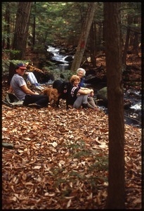 Dan and Nina Keller, with two dogs, mother and unidentified individual resting near stream in woods near Wendell Farm