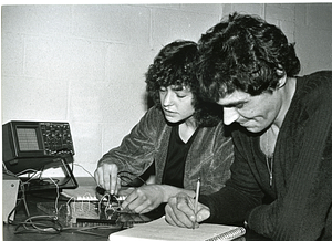 Unidentified woman working with electronics while an unidentified man takes notes