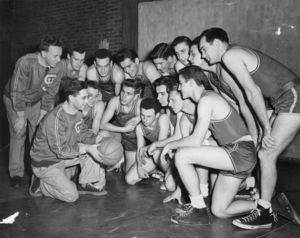 Suffolk University's basketball team crouched in a huddle, Coach Charlie Law in front holding ball, circa 1950s