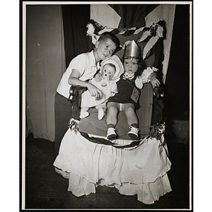 The Little Sister Contest winner sits on her throne with a baby doll while posing with her brother