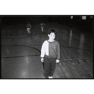 A smiling boy stands on an indoor court while two others run in the background