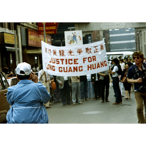Rear view of a man wearing a light blue jacket (in left foreground) taking a photograph of protesters holding a banner that reads, "Justice For Long Guang Huang" at a demonstration at the Chinatown gate
