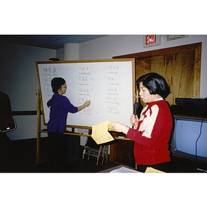 One woman reads from a sheet of paper while another woman writes the Association election results on a whiteboard