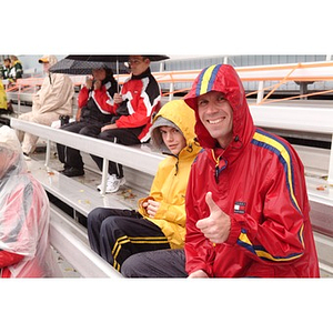 Two fans, dressed for rain, watch the game from the stands