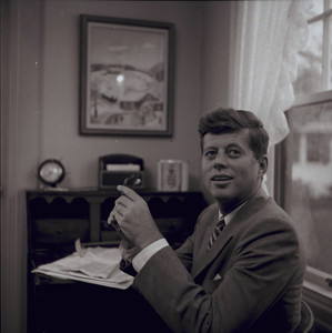 Portrait of John F. Kennedy, facing right, seated at his desk and holding eyeglasses, 1957
