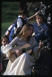 Judy Collins seated at the base of a statue, playing guitar for children