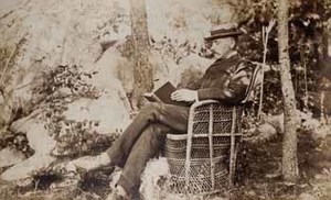 James Lowndes at Beverly Farms, seated outdoors in wicker chair, reading book, with dog at feet