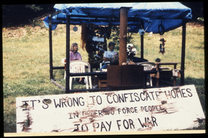 Supporters at the home of war tax resisters Randy Kehler and Betsy Corner seated behind a sign reading 'It's wrong to confiscate homes in order to force people to pay for war'