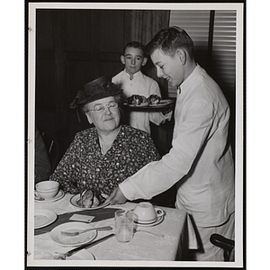 A boy dressed in a waiter's uniform serves a woman as another boy holding a tray looks on at a Mothers' Club event