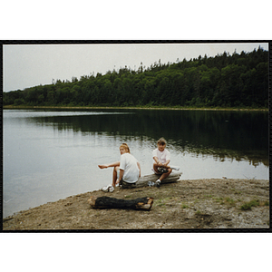 Two girls sit on a log on the shore of a pond