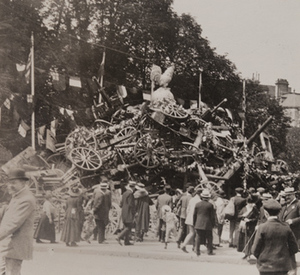 View of civilians in front of a large pile of captured German small artillery covered with wreathes, topped with a statue of an oversized rooster and flags from different countries hanging behind, 1919