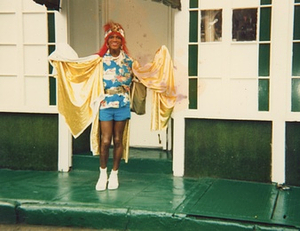 A Photograph of Marsha P. Johnson with Red Hair, Wearing a Blue Hawaiian Shirt and Draped in Yellow Fabric