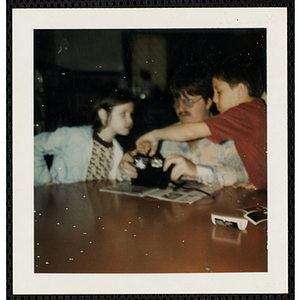 A man, a girl, and a boy from the South Boston Boys' Club sitting at a table, examining a camera