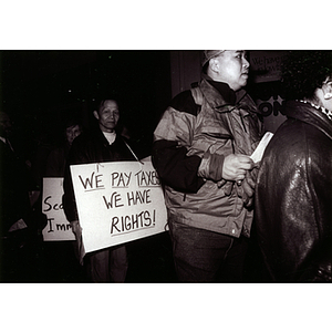 Demonstrators stand outside in Boston protesting for immigrant rights, with an Asian man holding a sign that reads, "We Pay Taxes, We Have Rights!"