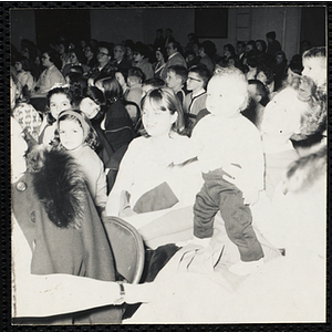 A toddler boy standing on a woman's lap to watch a show during a Boys' Club Week celebration event. A caption on the back of the photograph states "He likes the show, too"