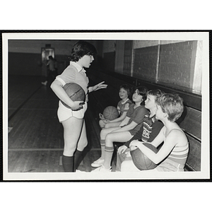 A girl holding a basketball speaks to four seated girls in a gym