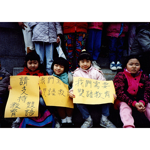 Four young girls sitting on the steps of the Massachusetts State House, holding protest signs in Chinese, at a rally for bilingual education in schools, with other children partially visible in the background